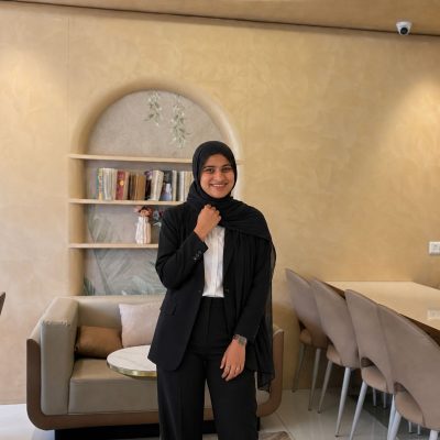 A smiling woman wearing a black hijab and black blazer suit with a white shirt, posing in a warm-toned café interior with arched shelving, books, and neutral-colored seating in the background.